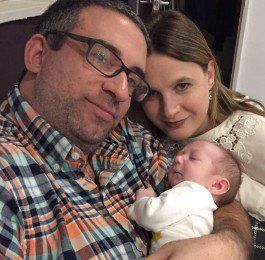 Two adults smiling close with a sleeping newborn in an adult’s arms