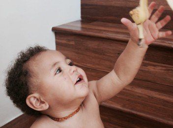 Toddler reaching up toward a stuffed toy on a wooden dresser, smiling in a room with carpeted stairs.