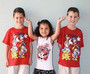 Three children standing side by side, smiling and raising their arms, wearing colorful graphic T-shirts.