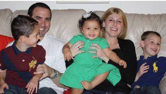 Family sitting on a couch, smiling with two children and a toddler in a green dress centered in the lap.