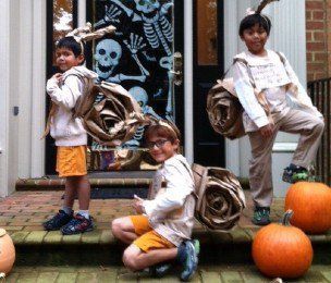 Three children in Halloween costumes posing on a porch with pumpkins and a skull decoration.