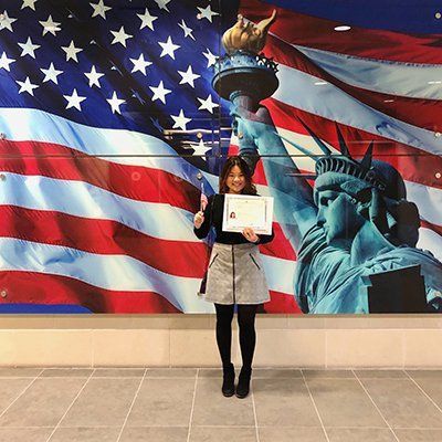 Person holding a certificate in front of a large U.S. flag and Statue of Liberty mural.