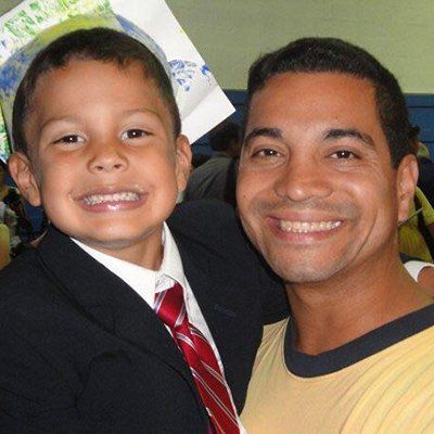 Smiling child in a suit poses beside a smiling man in a yellow shirt, both looking at the camera.