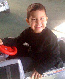 Smiling child in a black sweater riding a toy vehicle indoors, holding a red steering wheel