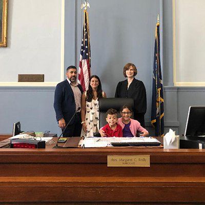 Family and official standing in a government chamber behind a wooden desk, smiling for a photo