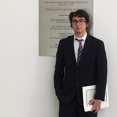 Man in a dark suit standing by a plaque, holding papers against a plain wall.