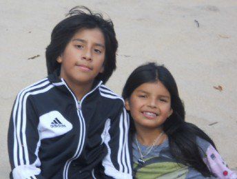 Two smiling children sitting together against a plain wall, one in a black Adidas jacket