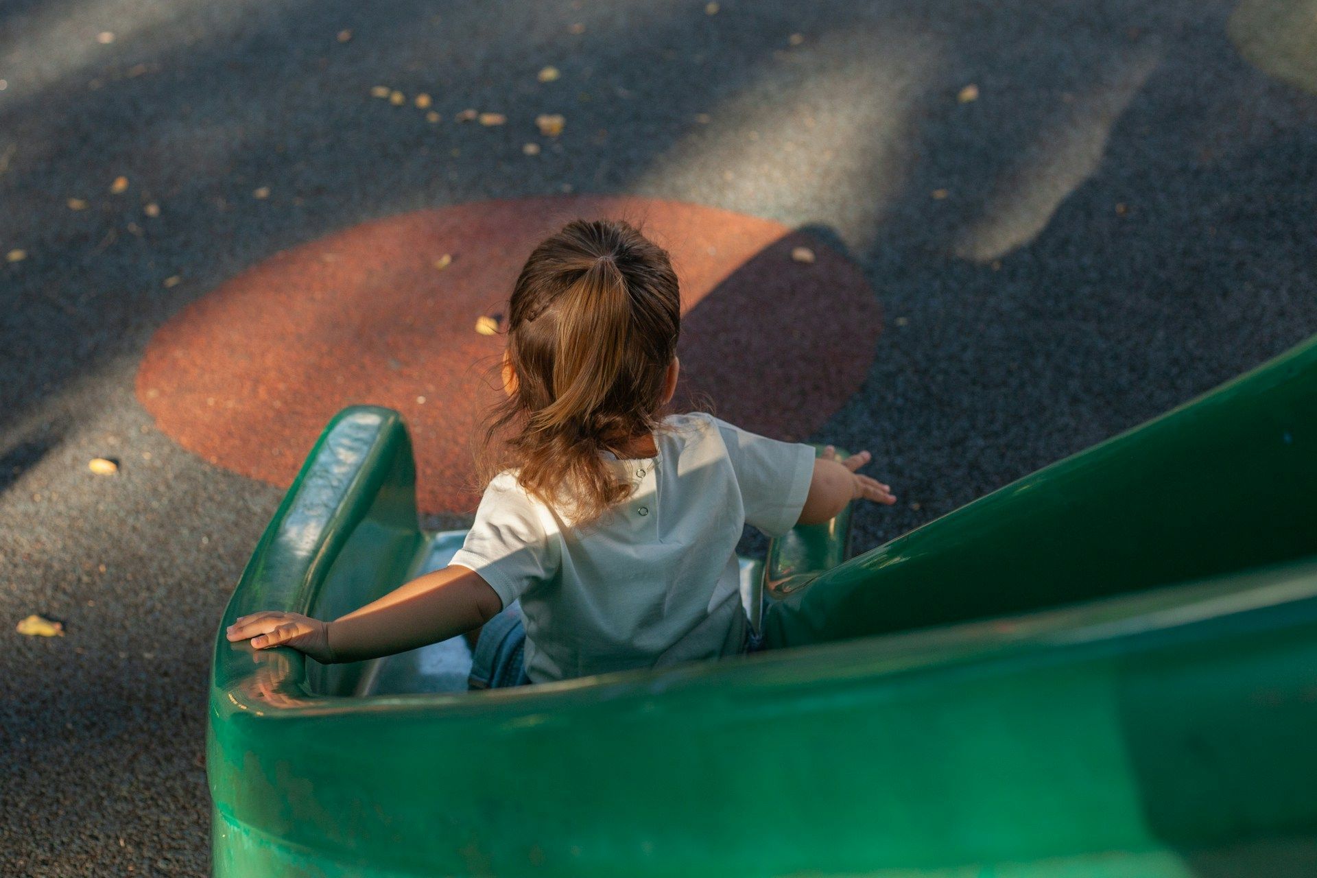 Child on a green slide at a playground, viewed from behind, with sunlit pavement below