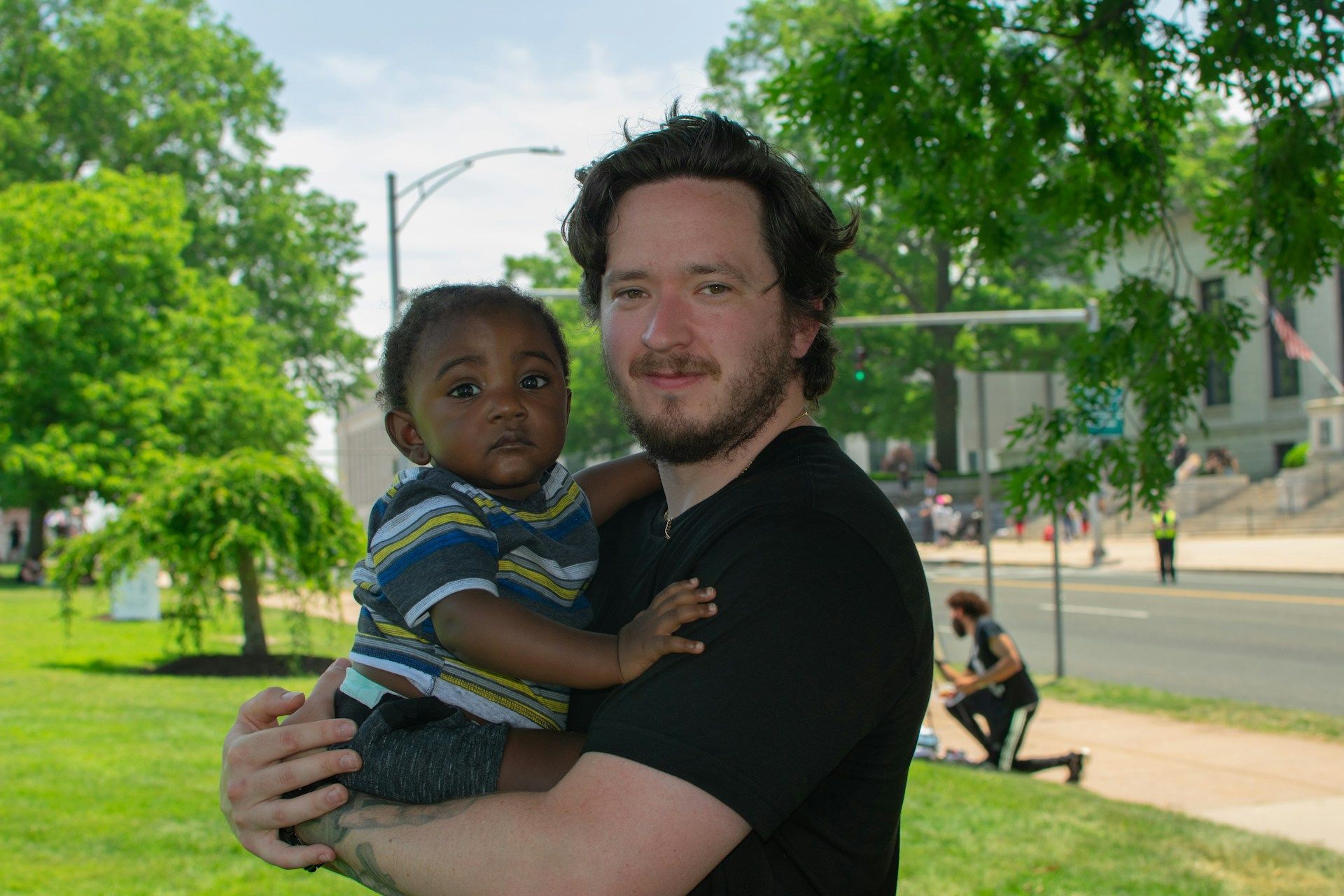 Man holding a child in a green park with trees and a sidewalk in the background