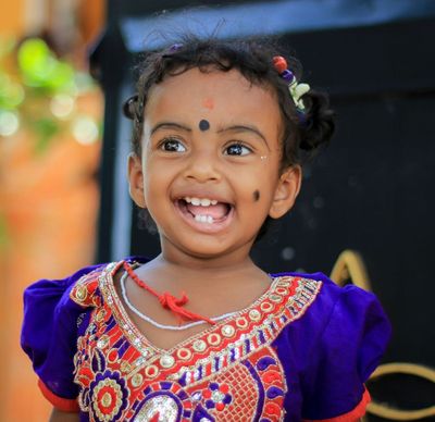 Smiling child in a purple embroidered outfit with a bindi, standing outdoors.