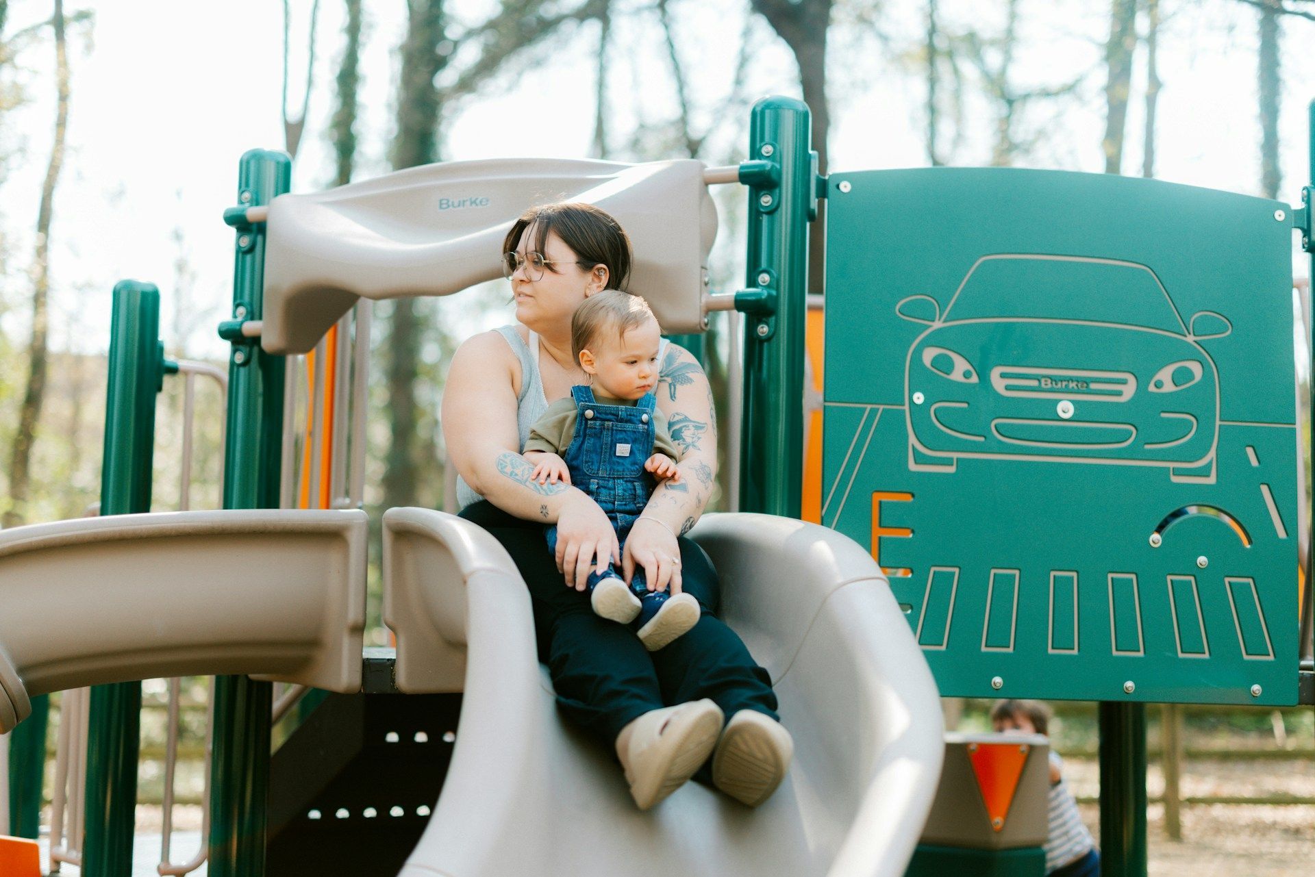 Adult holding a toddler on a playground slide beside a green climbing panel with a car design