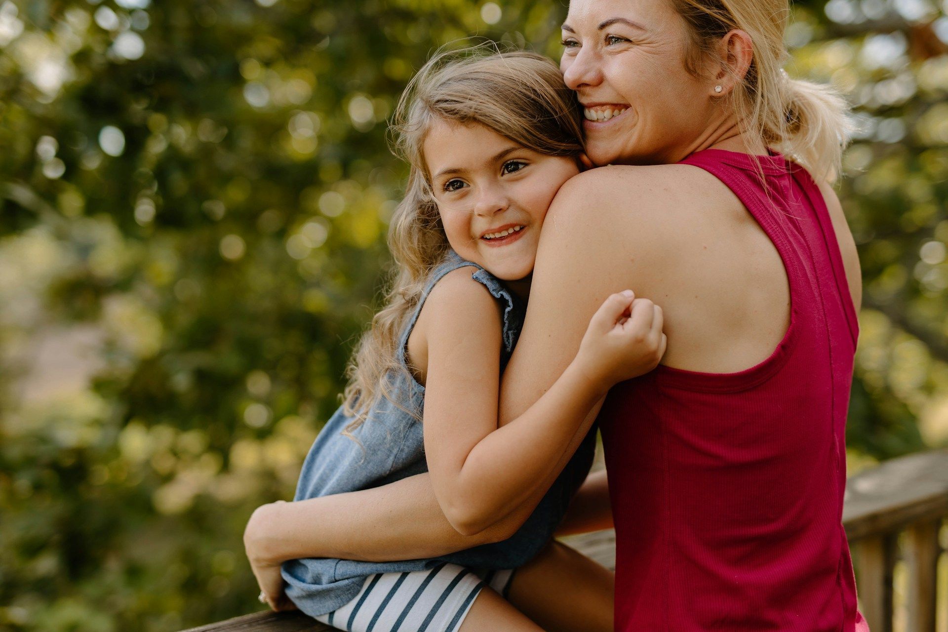 Woman in a red top hugging a smiling child outdoors with trees in the background