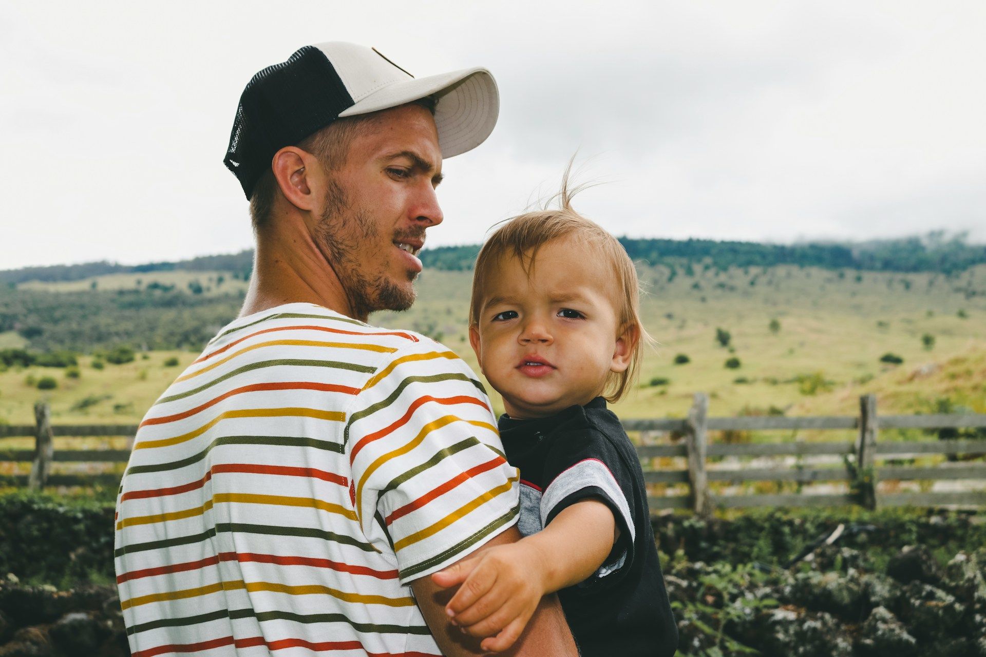 Man holding a toddler outdoors in a grassy field with hills and a wooden fence behind them