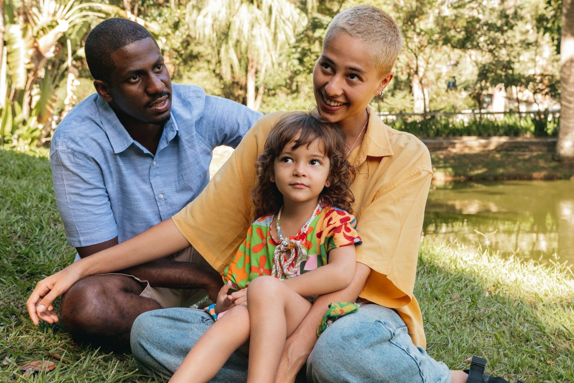 Three people sitting on grass by a pond, smiling in a sunny park