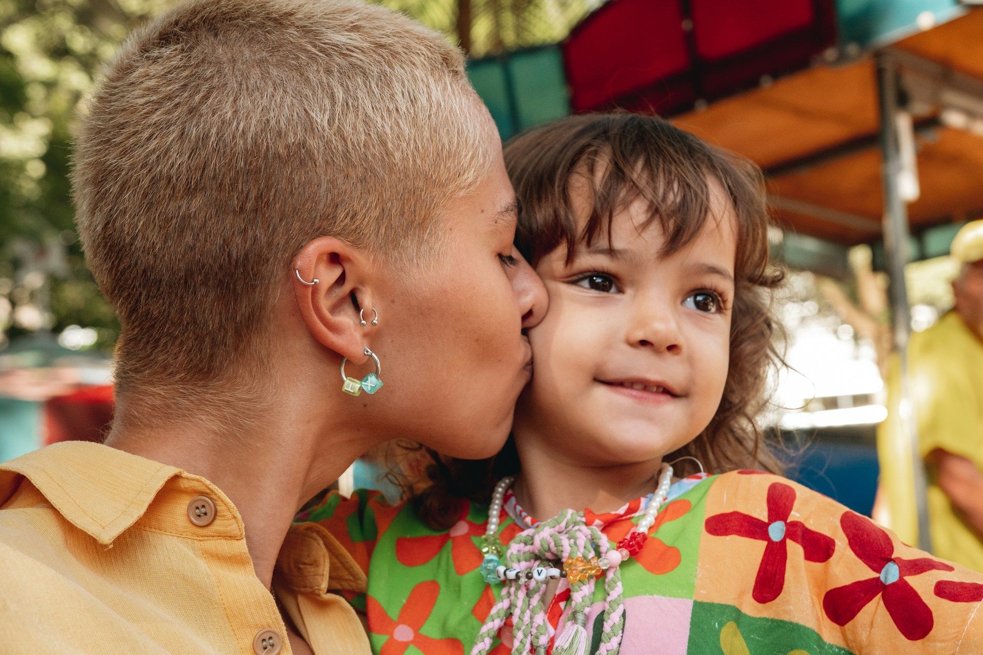 Person kissing a smiling child on the cheek, both in colorful patterned clothing at an outdoor setting