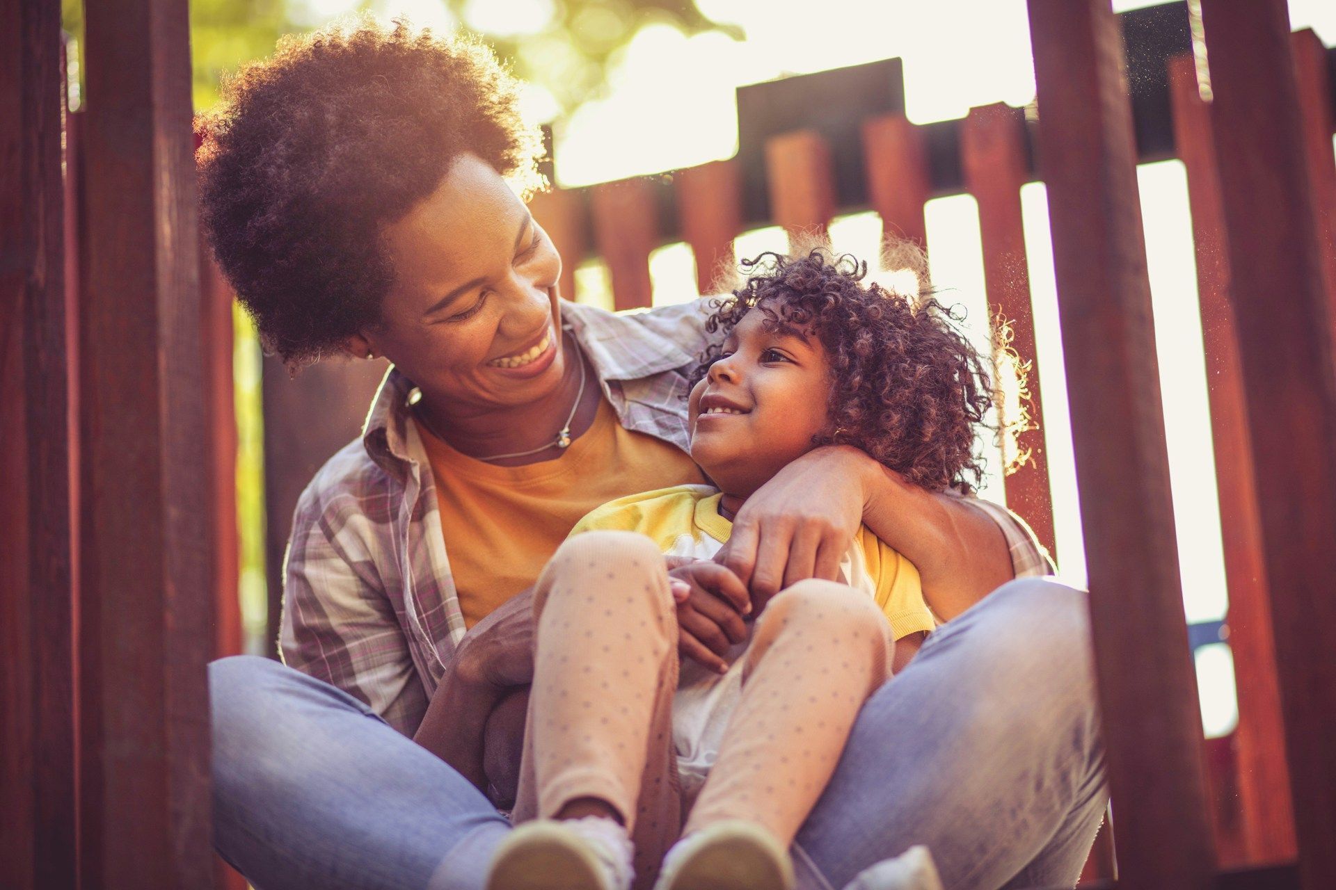 Adult smiling and holding a child on a porch, both in warm sunlight by a wooden fence.