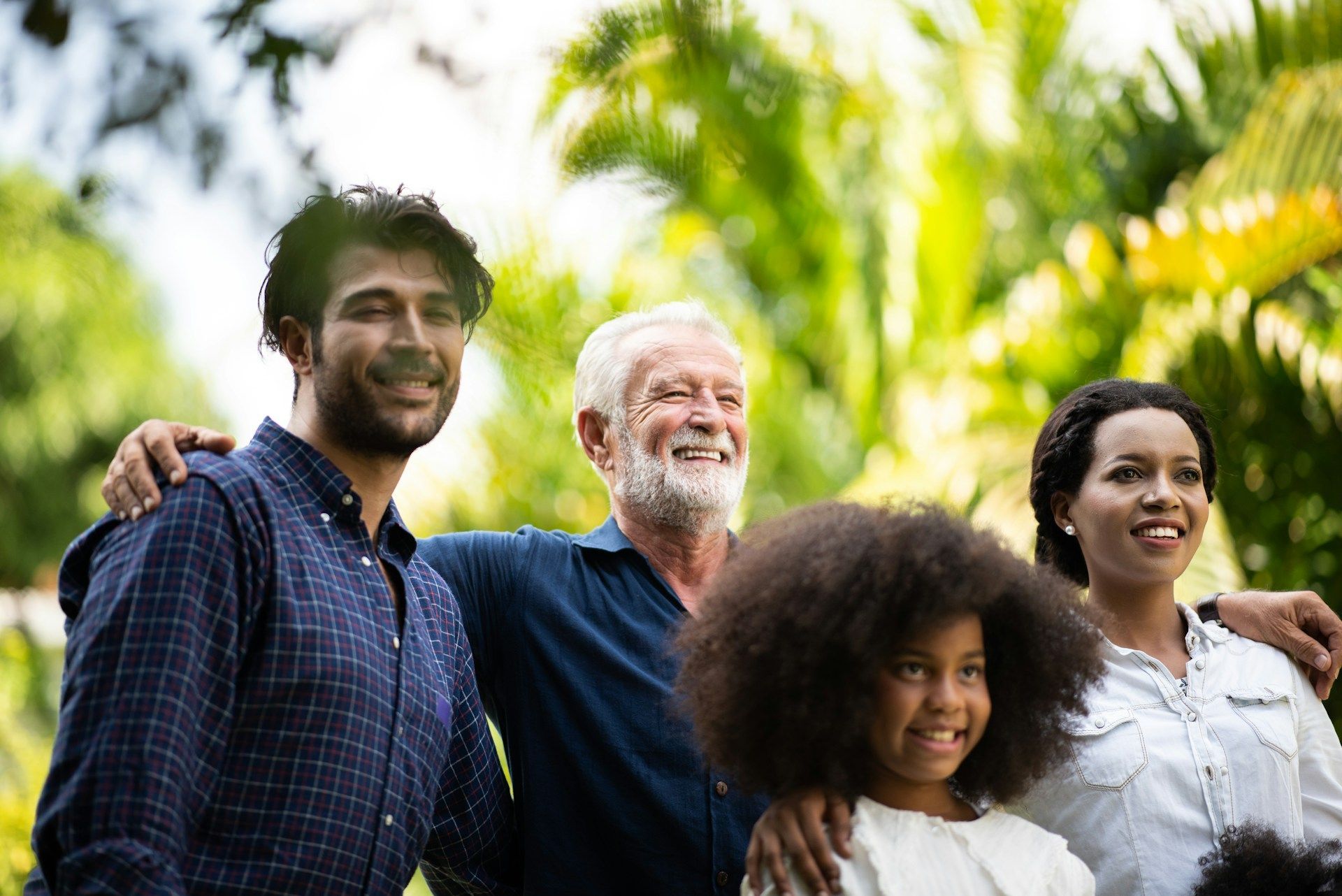 Four people smiling outdoors in a sunny park, with green trees in the background.