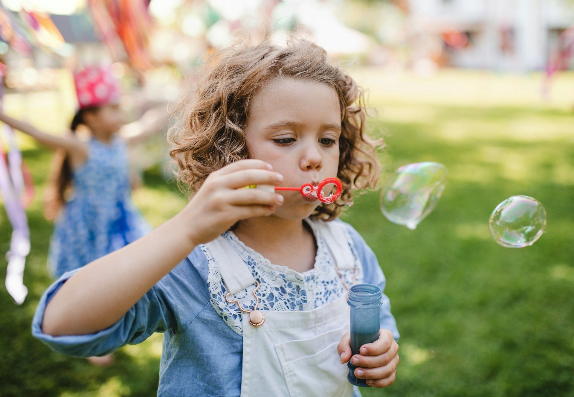 Child blowing bubbles in a sunny park, holding a bubble bottle with soap bubbles floating nearby.
