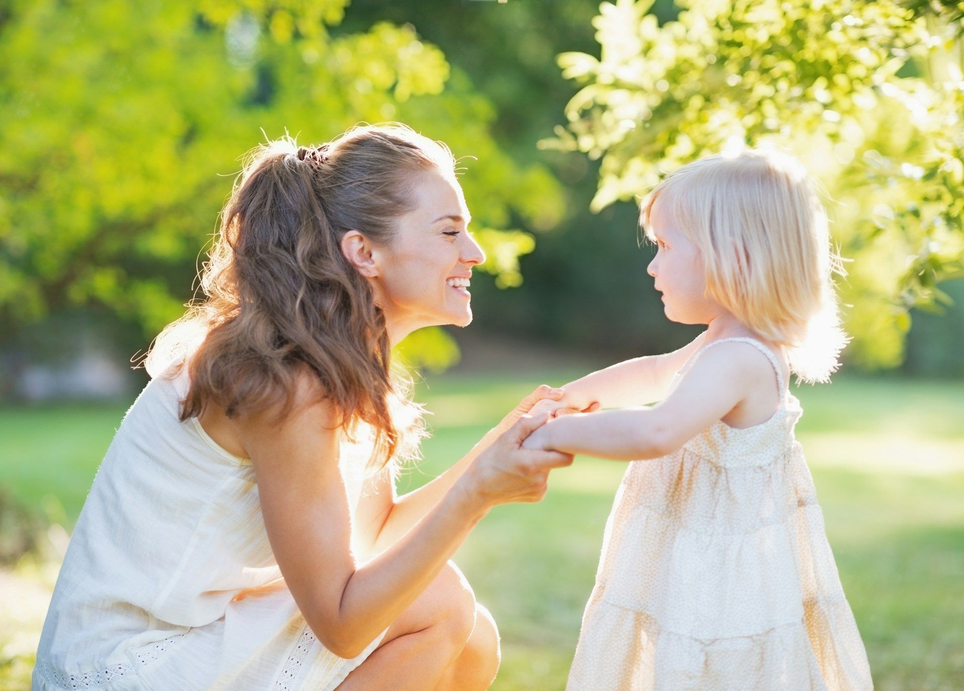Woman kneels smiling at a child in a white dress in a sunlit park, holding hands.