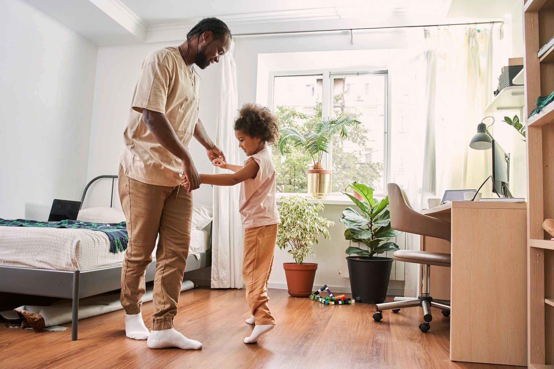 Adult and child holding hands in a bright home office with a bed, plants, and wooden desk