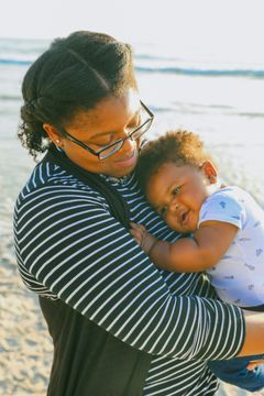 Person holding a sleeping baby outdoors near water, both in striped clothing, with warm sunlight.