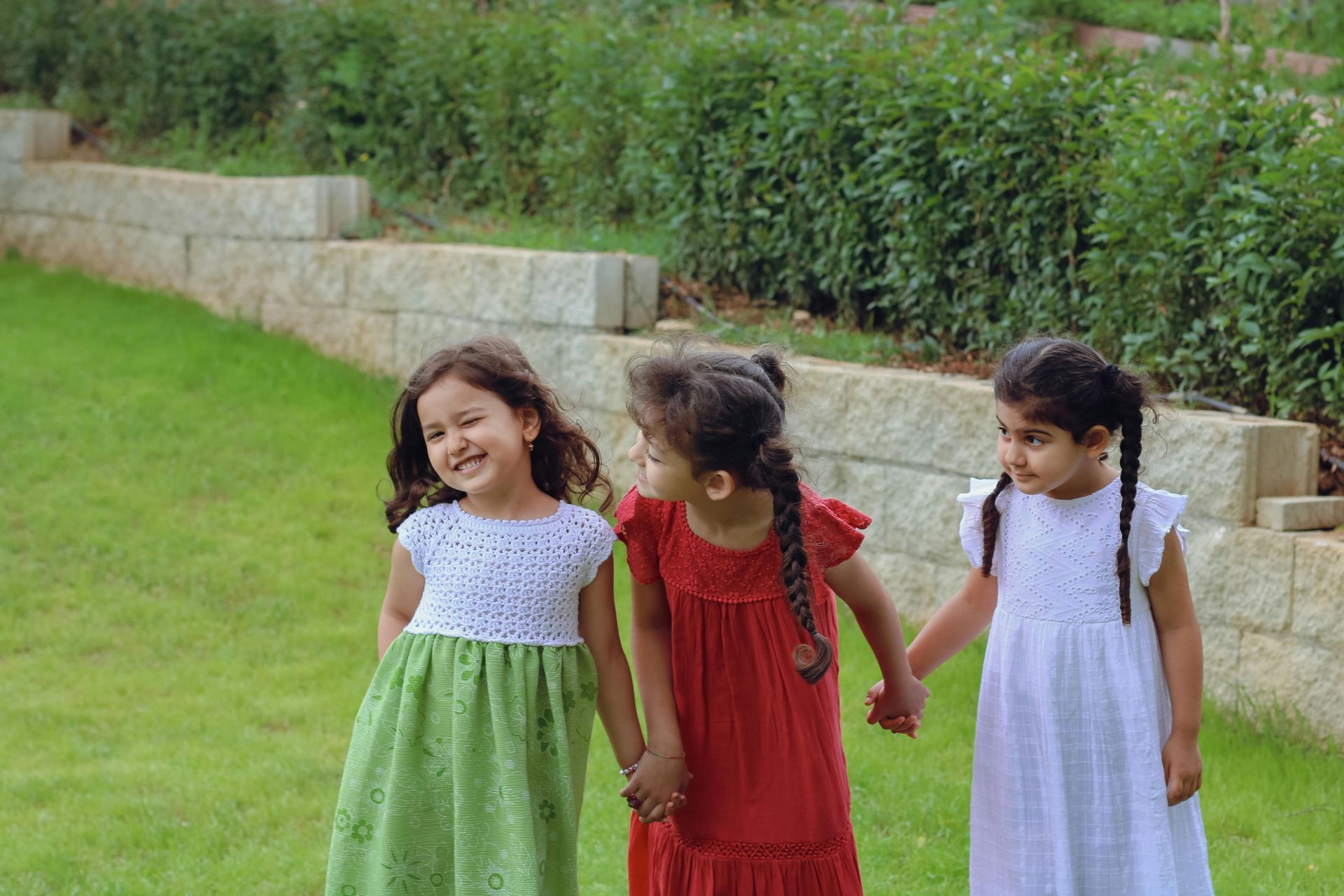 Three children holding hands and smiling while walking on grass near stone steps and shrubs.