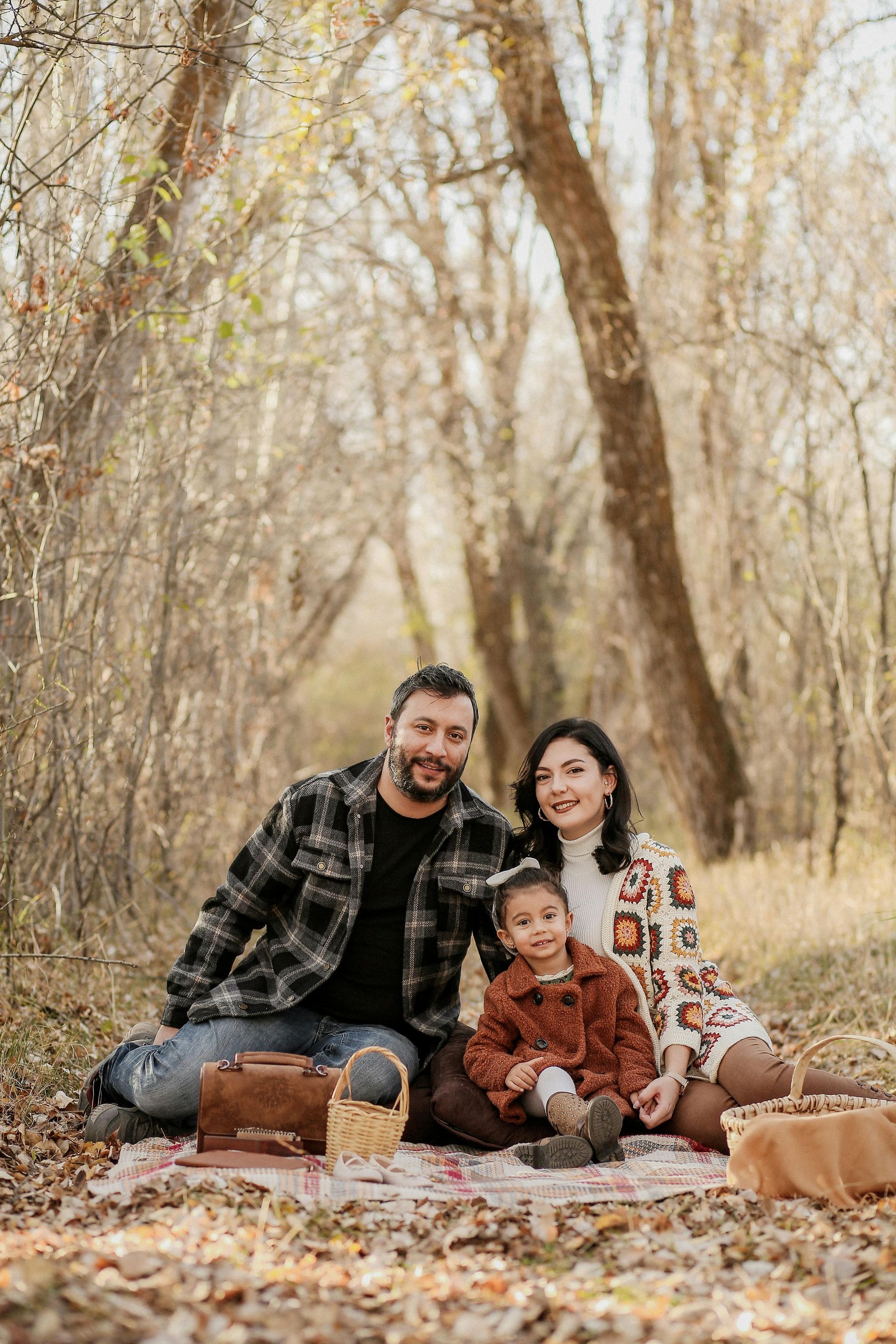 Family sitting together on a leaf-covered path in a wooded autumn setting, smiling at the camera