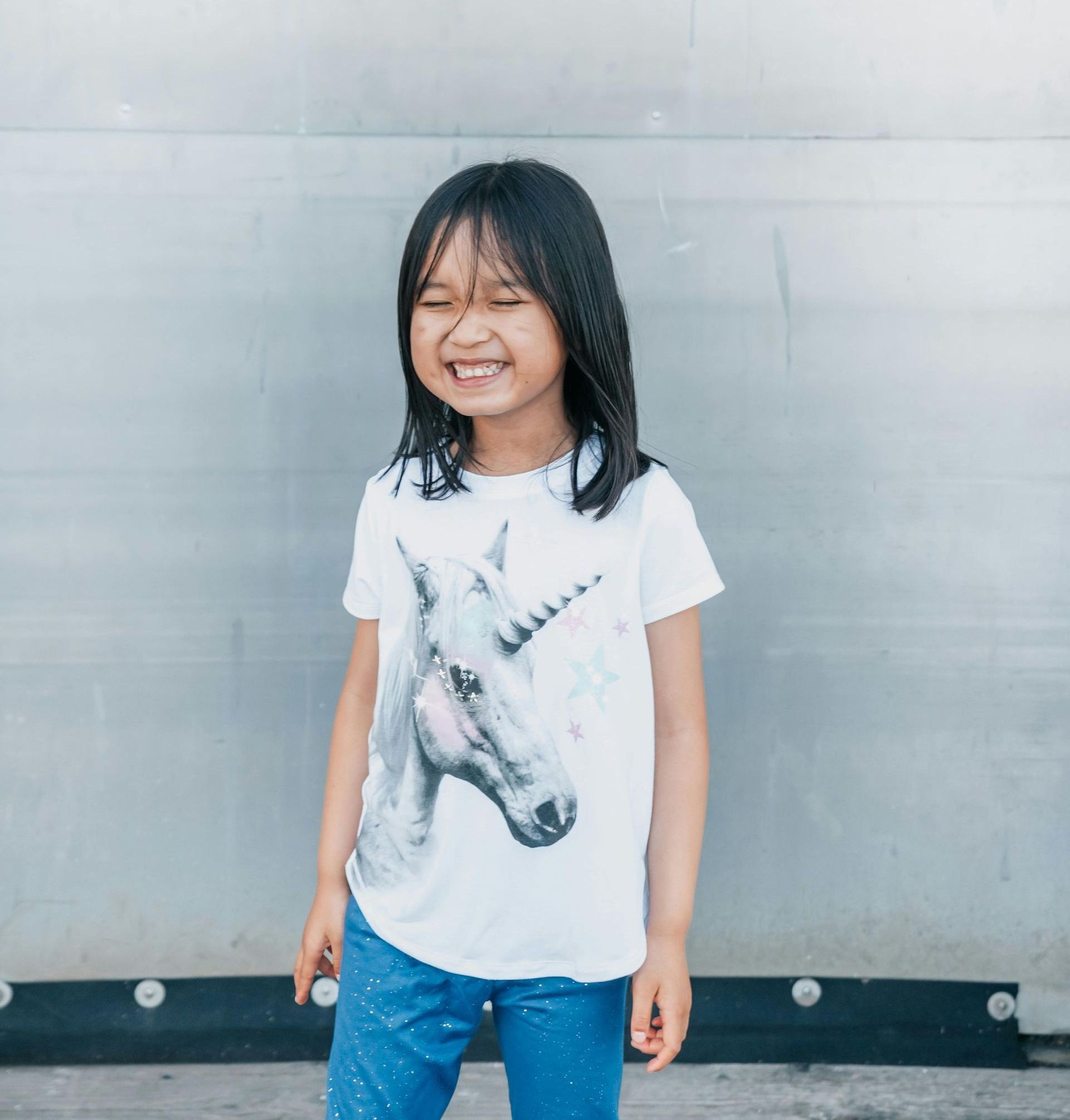 Smiling child in a white unicorn T-shirt and blue pants standing against a gray wall