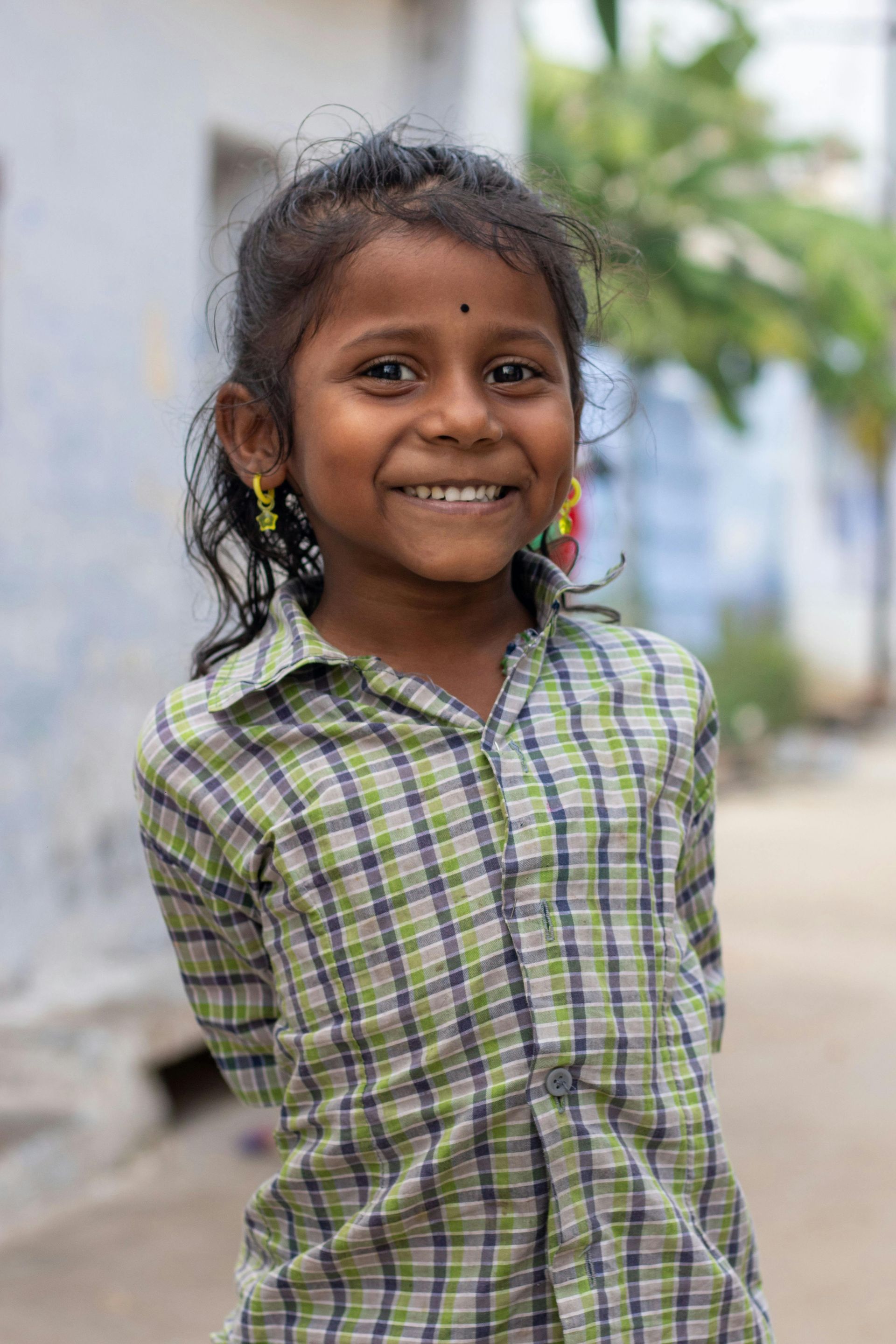 Smiling child in a green plaid shirt standing outdoors on a blurred street.