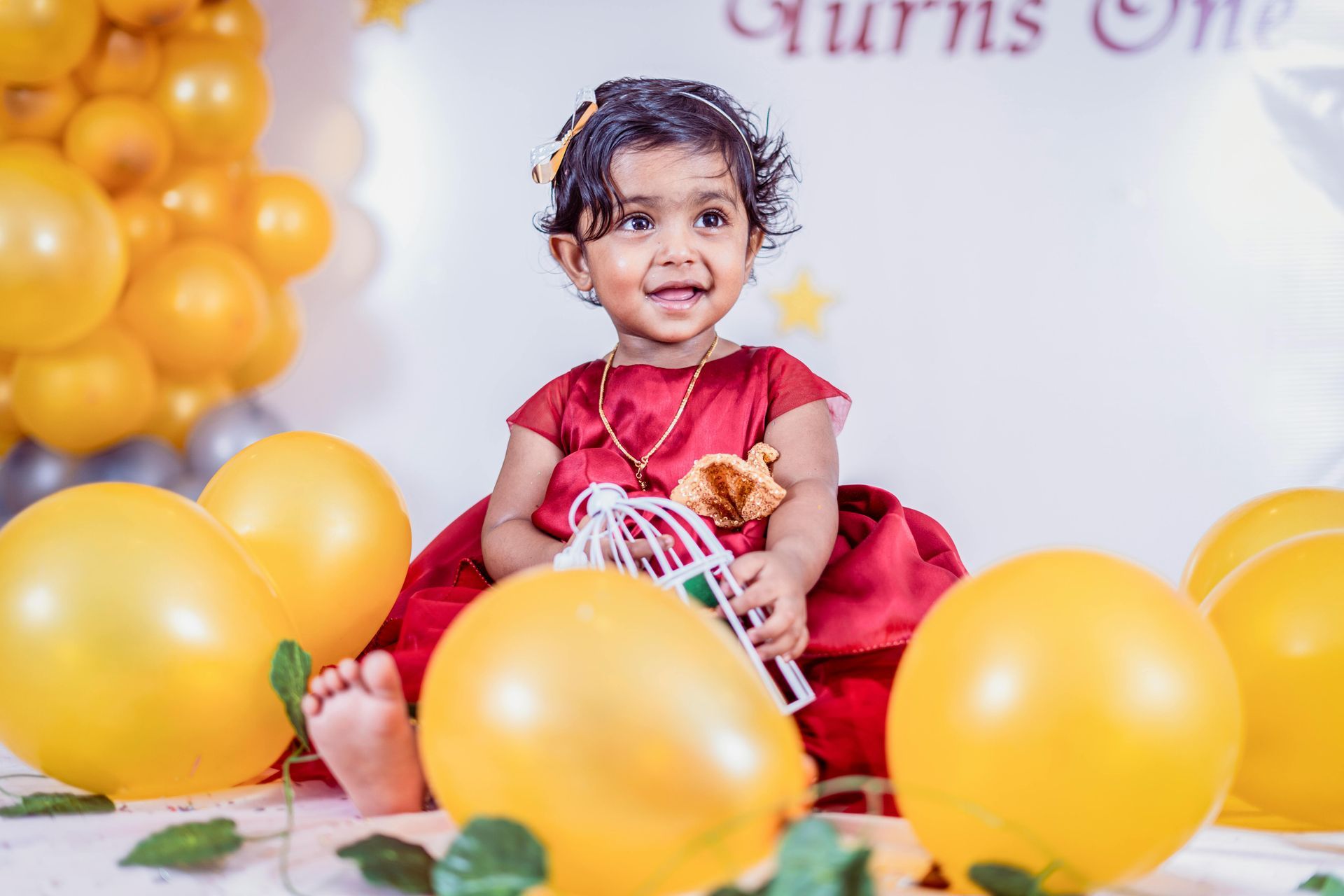 Toddler sitting among yellow balloons, wearing a red dress and smiling at a birthday party.