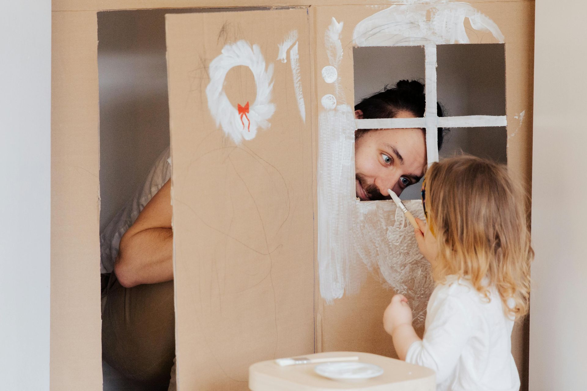 Man peeking through cardboard playhouse windows at a child inside a cardboard fort