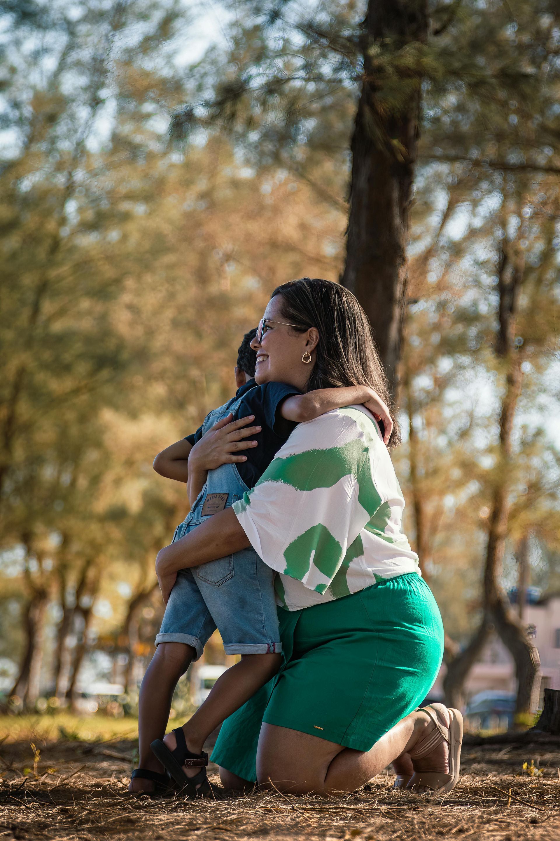 Two people hugging outdoors in a park, one kneeling in green shorts and striped shirt, holding a child in denim shorts.