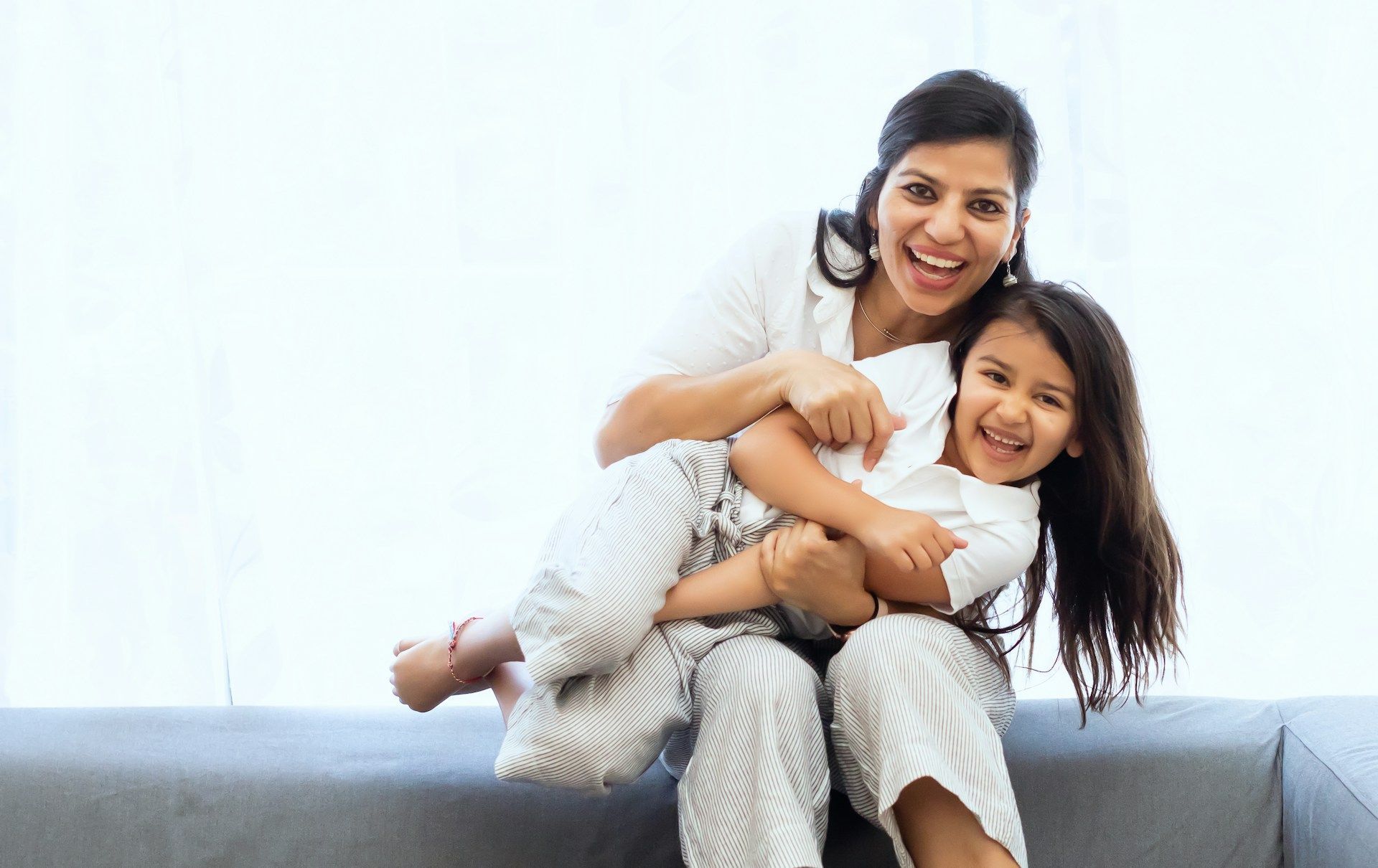 Two smiling people sitting on a couch, hugging and laughing in a bright room.