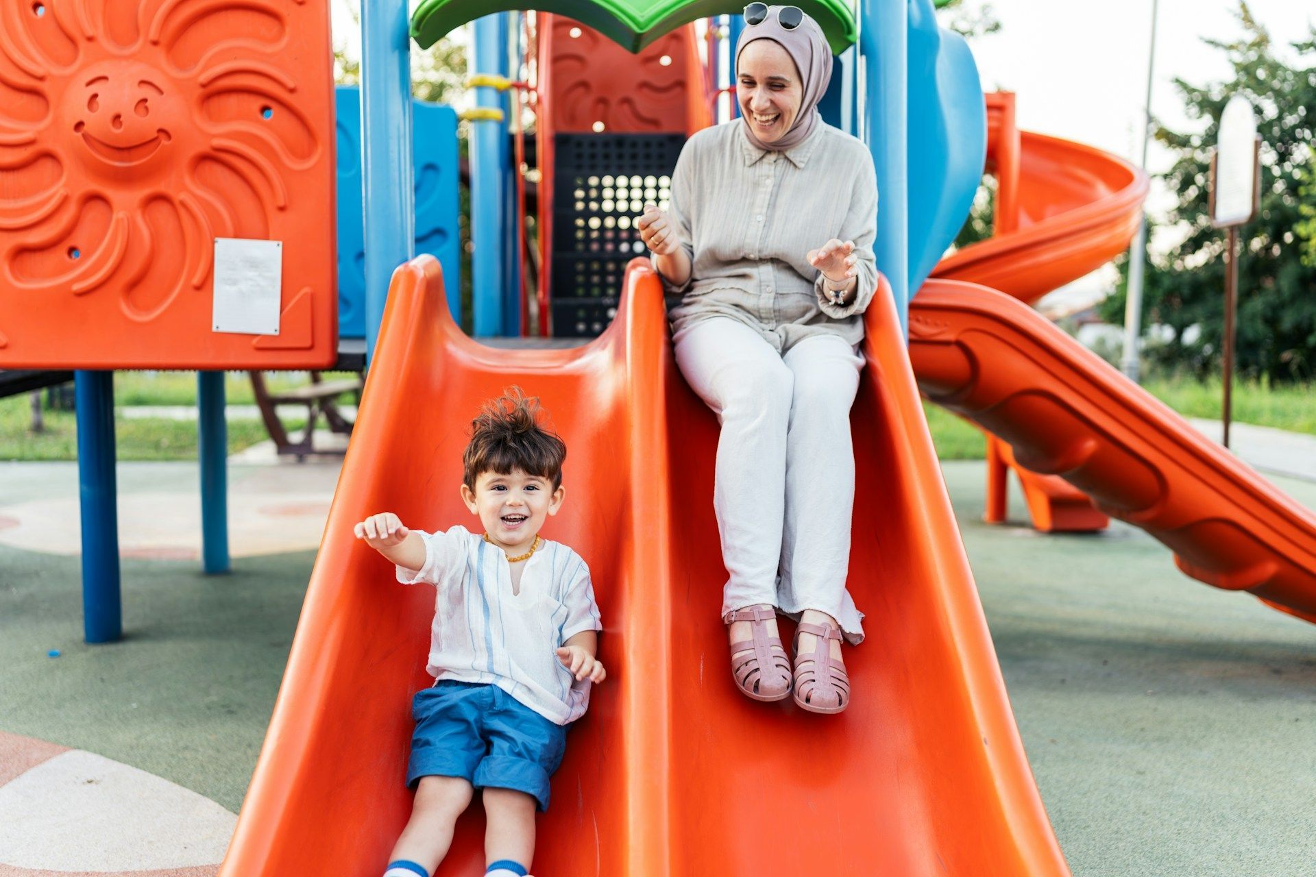 Woman and child sitting on a bright orange playground slide, smiling outdoors.