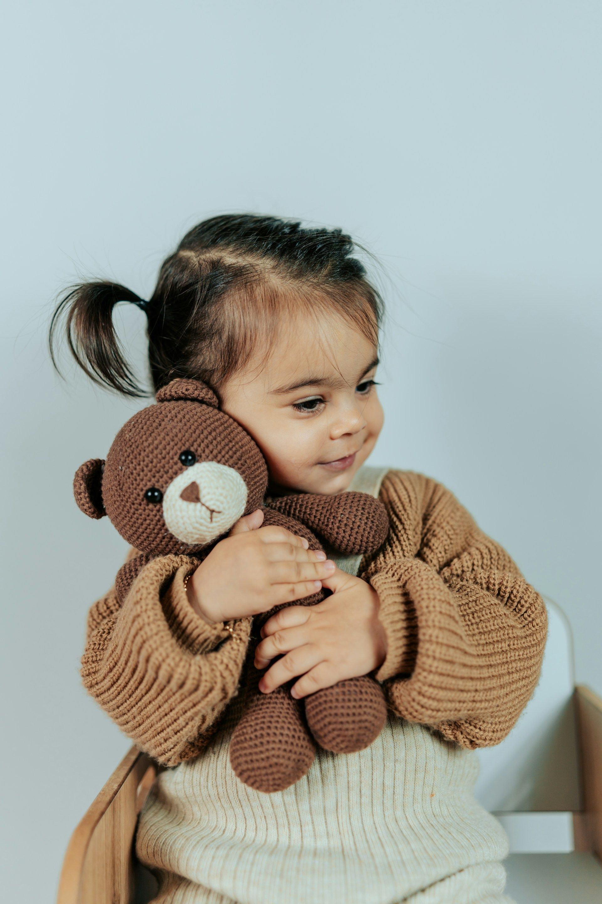 Child hugging a brown teddy bear, wearing a tan sweater against a light background.