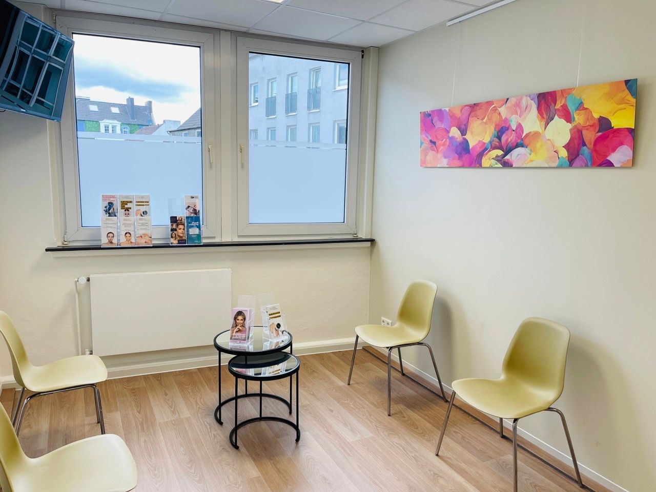 Wartezimmer mit beigen Stühlen, Kunstwerken, kleinen Tischen und einem Fenster.