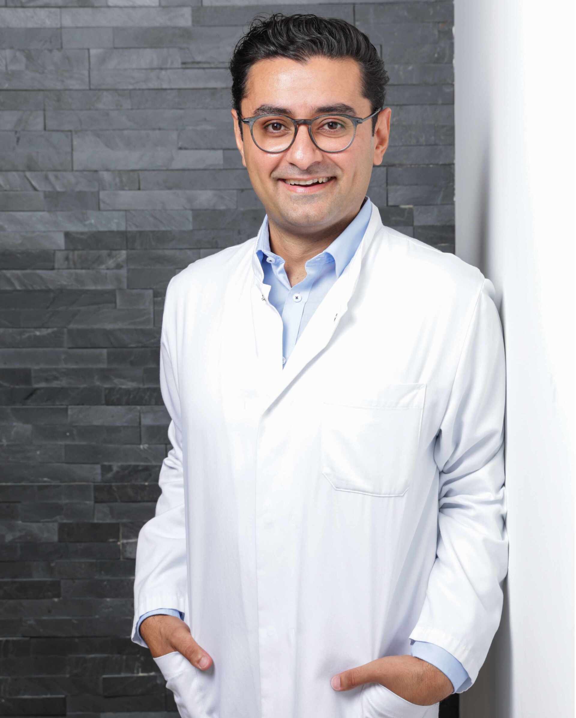 Dr. Hessam im Kittel lächelt und lehnt an einer grauen Backsteinmauer. Er trägt eine Brille und ein hellblaues Hemd.
