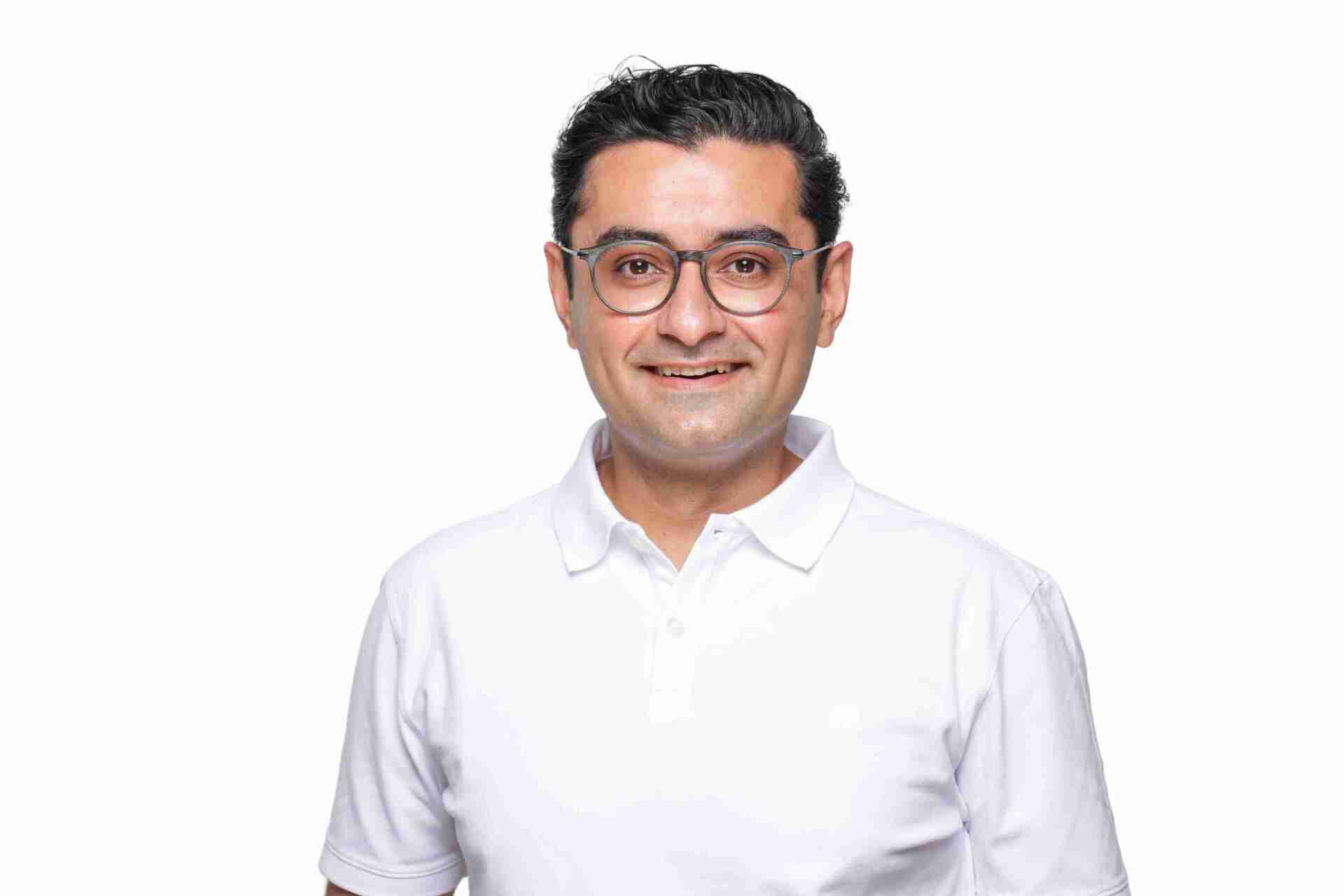 Dr. Hessam mit Brille lächelt, weißes Poloshirt, vor weißem Hintergrund.