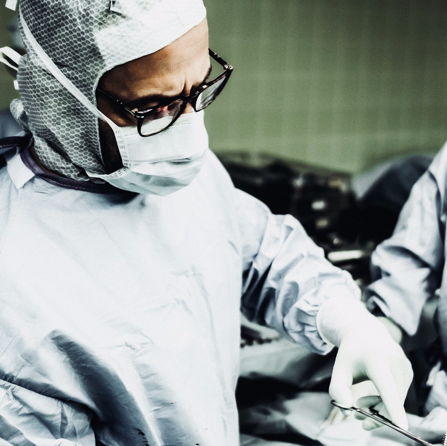 Chirurg in steriler Kleidung, mit Brille, hält eine chirurgische Schere in einem Operationssaal.