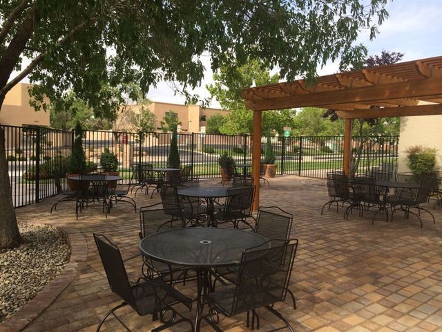 Outdoor patio with black tables and chairs under a wooden pergola and trees, on a brick-patterned surface.