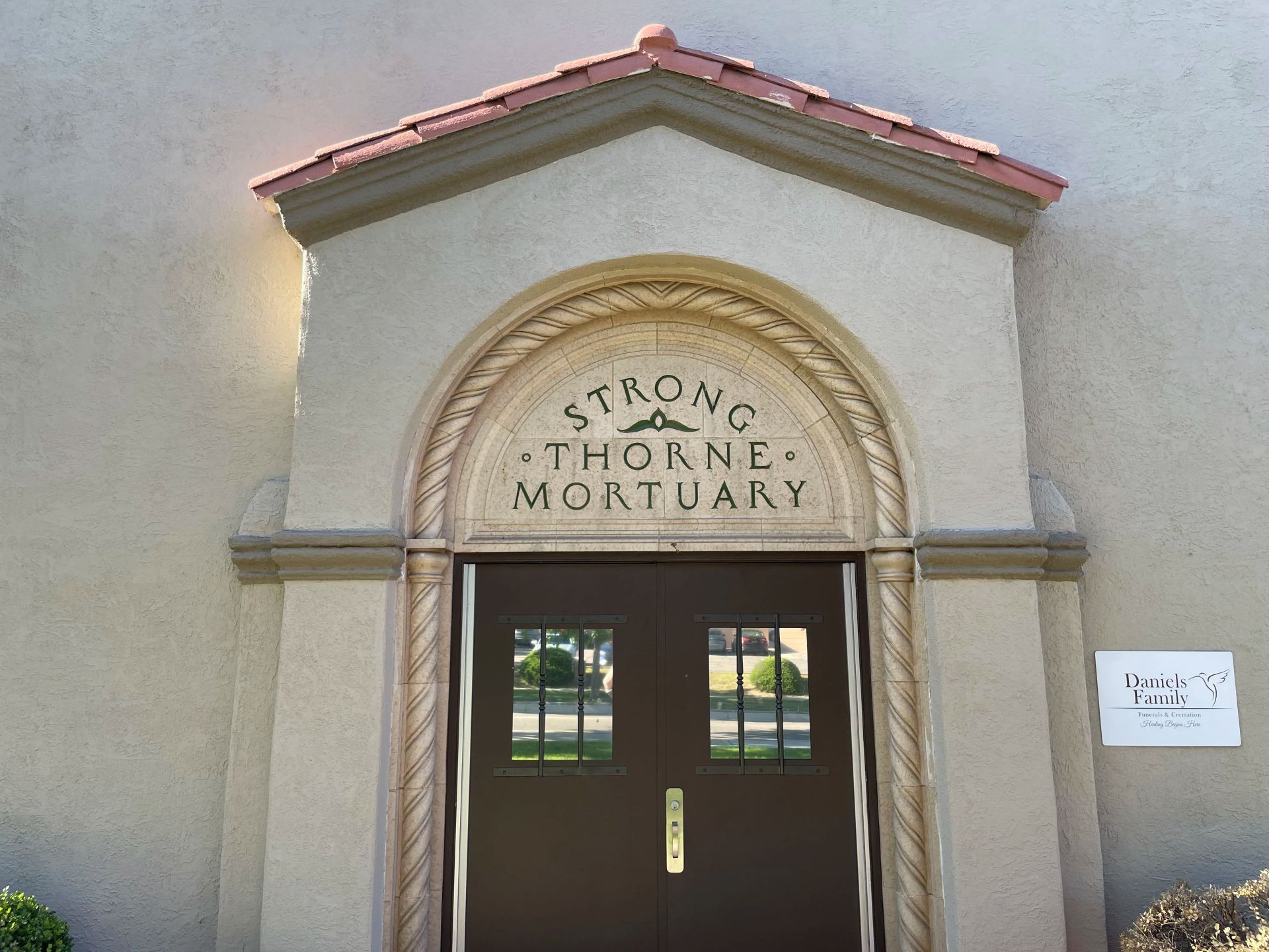 Entrance to Strong-Thorne Mortuary. Beige stucco building with brown double doors. Decorative stone archway with the name.