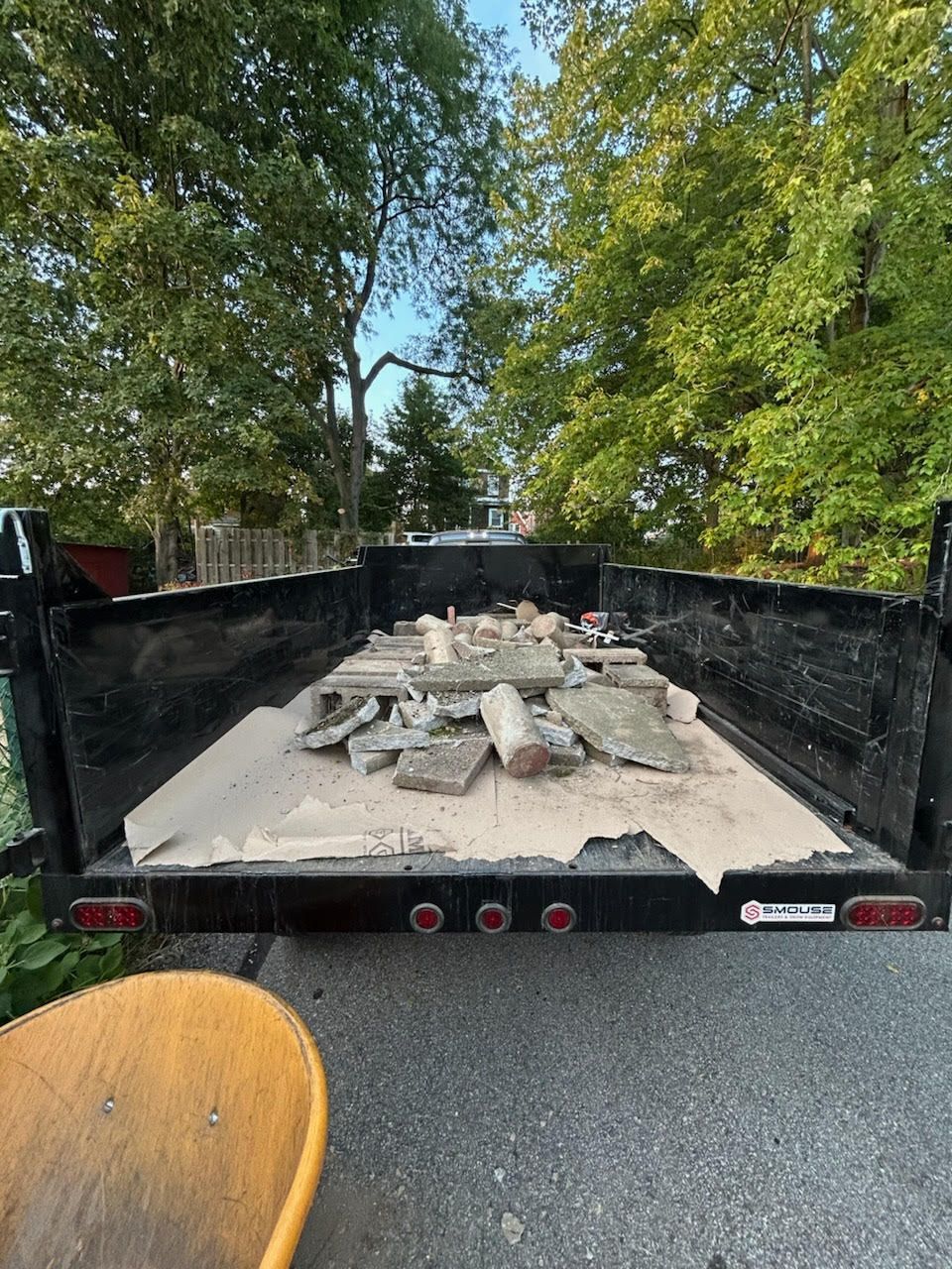 A black trailer bed filled with concrete debris, parked on a paved surface near trees and a wheelbarrow.