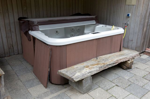Hot tub with brown siding and cover next to a bench on a brick patio.