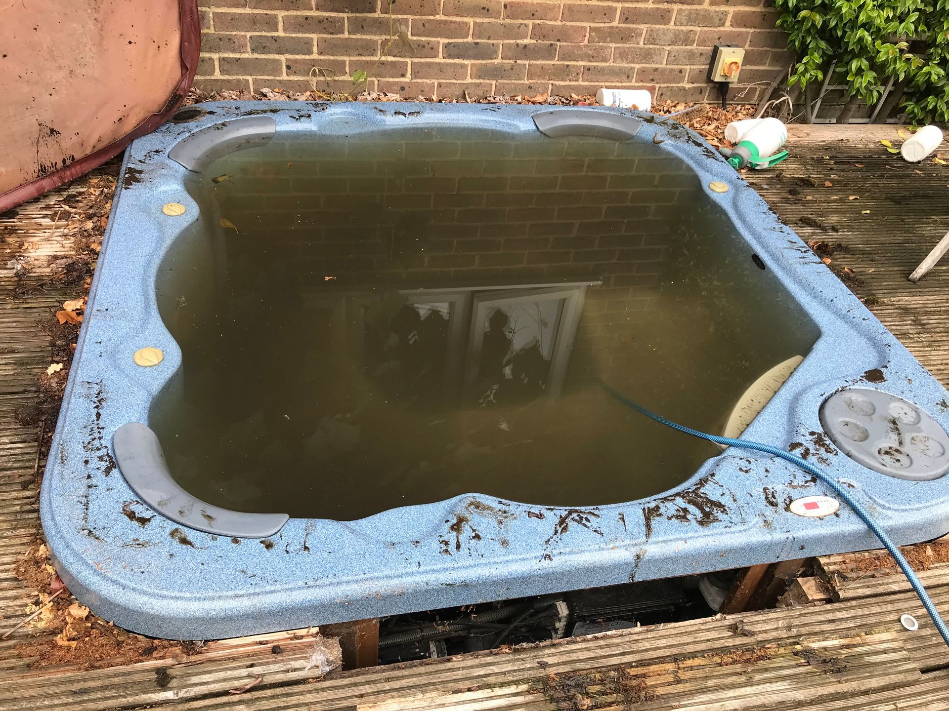 Dirty, algae-filled hot tub on a wooden deck, surrounded by debris and a brick wall background.