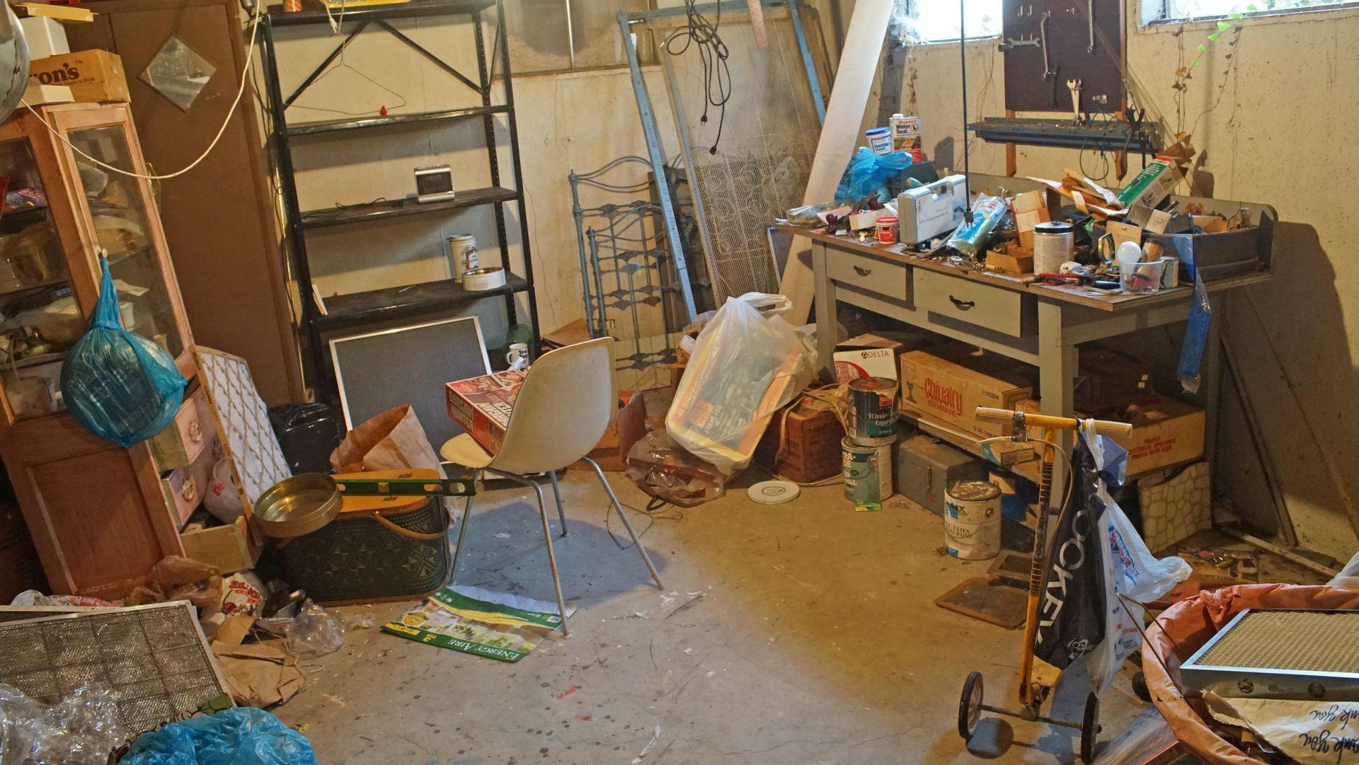 Cluttered basement with shelves, table, chair, trash bags, and miscellaneous items.