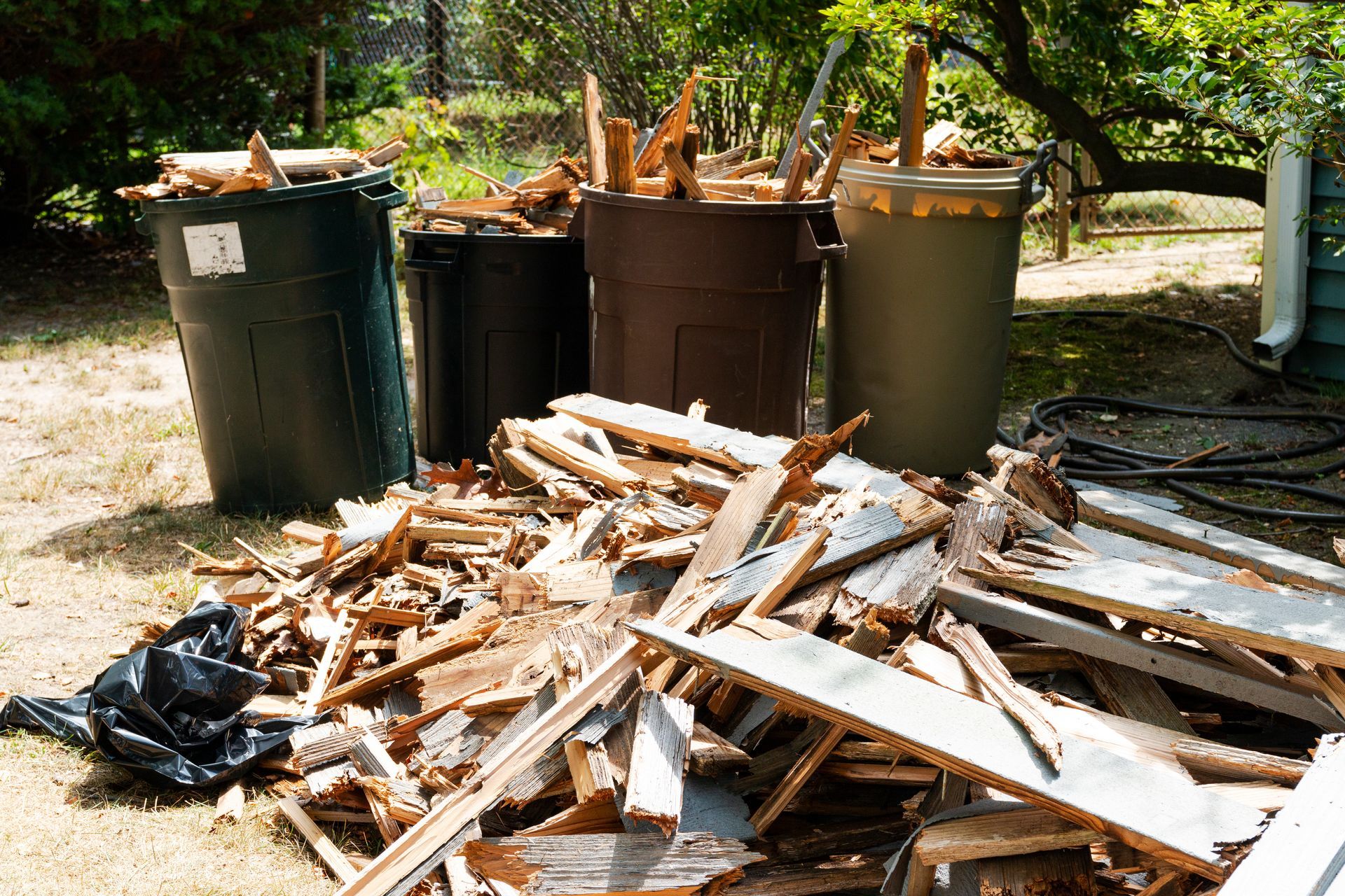 Debris pile with overflowing trash cans in a yard; wood, green and brown containers.