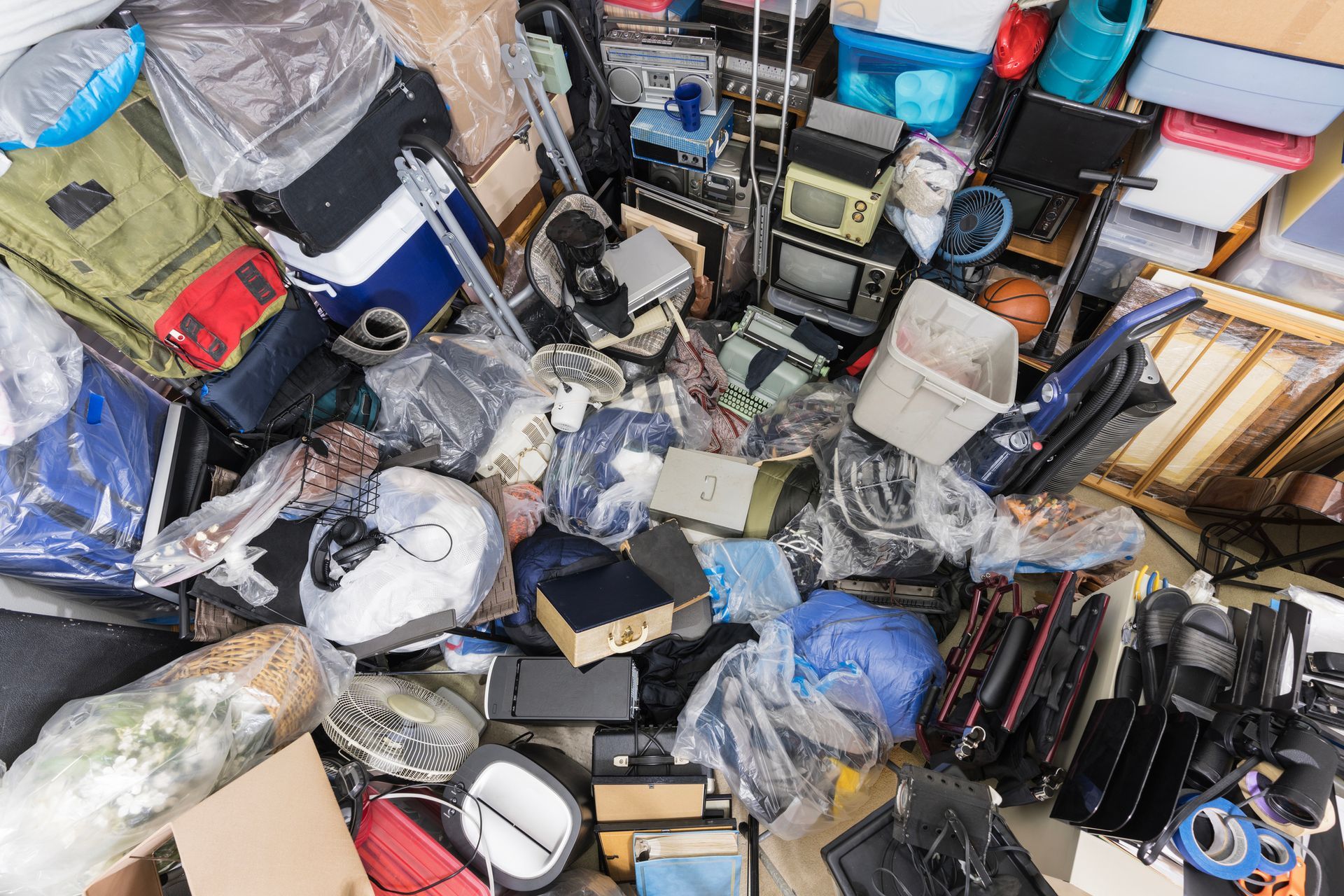 Messy room, overflowing with various items including luggage, electronics, boxes, and plastic wrapping.