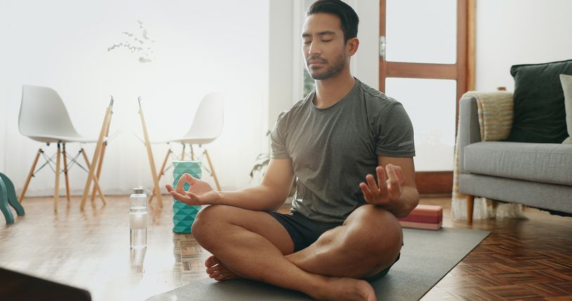 Man meditating in lotus position on a yoga mat in his living room.