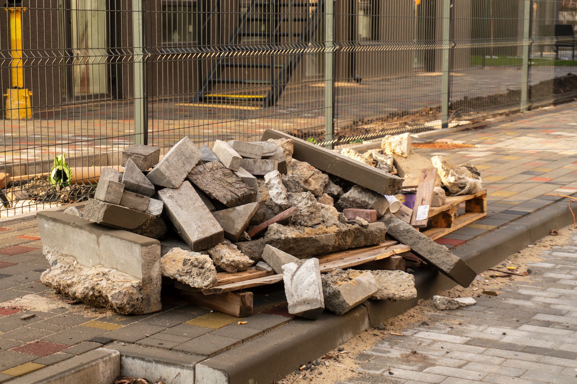 Pile of rubble on a wooden pallet next to a curb and sidewalk, with a fence in the background.