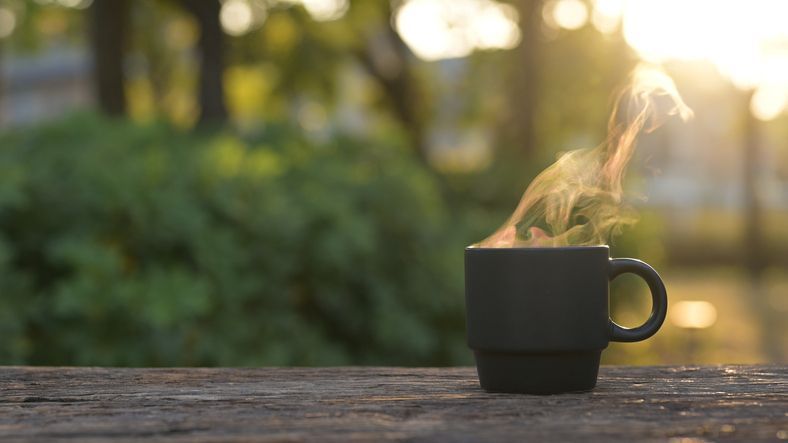Steaming black mug on a wooden surface, sunlit green background.
